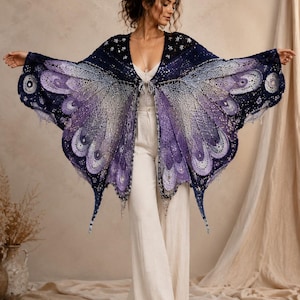 May include: A crochet shawl featuring a butterfly wing pattern in shades of deep navy, violet, and light lavender. The garment has an open front with a tie closure, intricate lace-like stitching, and scalloped edges that mimic the shape of wings.