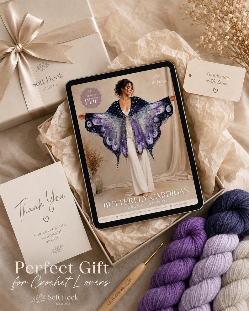 May include: A digital tablet displaying a crochet pattern for a butterfly cardigan, accompanied by three skeins of yarn in shades of purple, a wooden crochet hook, a gift box with a ribbon, and a thank you card from Soft Hook Studio.