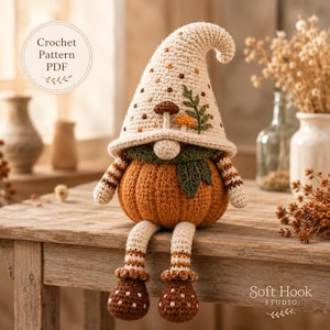 Può includere: Una bambola gnomo all'uncinetto con un corpo arancione a forma di zucca, lunghe gambe penzolanti e un cappello a punta color crema decorato con piccoli punti marroni, una foglia verde e due piccoli funghi. Il testo Crochet Pattern PDF è visibile.