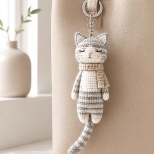 May include: A crochet cat keychain featuring a grey and white striped body, a beige scarf, and a silver metal ring. The cat has embroidered closed eyes, a small pink nose, and whiskers. It is attached to a beige leather-like handbag.