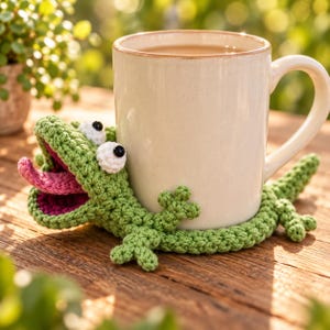 Puede incluir: Una taza de cerámica de color beige claro sobre un posavasos de ganchillo verde con forma de lagarto. El lagarto tiene la boca muy abierta con una lengua rosa, dos ojos redondos blancos con pupilas negras y una cola larga alrededor.