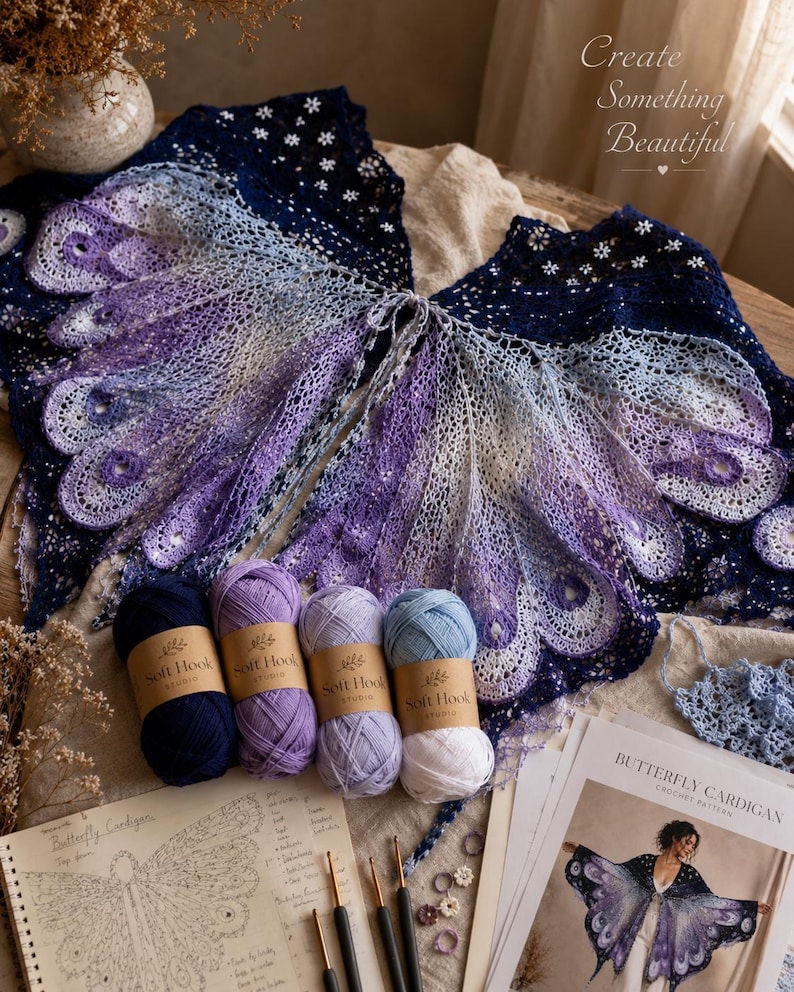 May include: A crochet butterfly cardigan featuring a gradient of blue, purple, and white yarn, displayed with four skeins of yarn, crochet hooks, stitch markers, and a printed pattern titled Butterfly Cardigan with the text Create Something Beautiful.