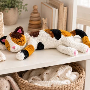 May include: A crocheted calico cat plush toy featuring a white body with patches of black and orange yarn. The cat has a sleeping expression with embroidered closed eyes, a pink nose, and pink inner ears, with a long tail extended outward.