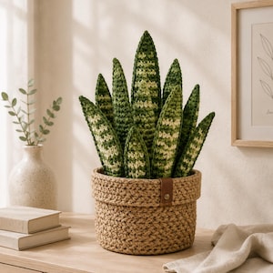 May include: A crochet snake plant with variegated dark and light green leaves, potted in a textured, woven tan basket featuring a small brown leather tab with a metal rivet. The plant has multiple upright, pointed leaves with a distinct striped pattern.
