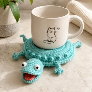 May include: A light blue crocheted coaster shaped like a lizard with a textured back, white eyes, and a pink tongue, supporting a white ceramic mug featuring a black line drawing of a sitting cat and a small heart symbol on its front surface.
