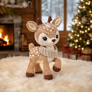 Puede incluir: Un peluche de cervatillo tejido a ganchillo con pelaje marrón claro, manchas blancas en el lomo y la cabeza, pezuñas marrón oscuro y astas pequeñas. El cervatillo lleva una bufanda de ganchillo gris claro texturizada y tiene grandes ojos negros.