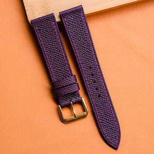 Może przedstawiać: Para głębokofioletowych pasków do zegarka z różowymi przeszyciami i złotą klamrą. Paski wykonane są z teksturowanej skóry i są prezentowane na pomarańczowej powierzchni.