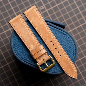 Pode incluir: Uma pulseira de relógio em couro castanho-claro com costuras claras, uma fivela de metal dourado e dois passadores de couro. A pulseira está sobre um objeto oval azul texturizado com um padrão de grão visível e bordas costuradas.