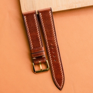 May include: Two brown leather watch straps with white stitching and gold-colored buckles. The straps are made of a rich, reddish-brown leather and are displayed on a light orange surface.