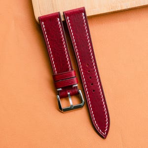 Puede incluir: Correas de reloj de cuero burdeos con costuras blancas y hebilla plateada. Las correas se muestran sobre una superficie naranja con un elemento de madera en el fondo. Las correas son nuevas.