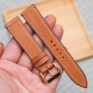 Puede incluir: Un par de correas de reloj de cuero marrón claro con costuras blancas. Las correas tienen una hebilla dorada y están sujetas en una mano. Las correas del reloj son de cuero texturizado.