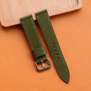 以下が含まれることがあります： オリーブグリーンの革製腕時計ストラップ。白いステッチとシルバーのバックルが付いています。オレンジ色の表面に、背景に木製のトレイを置いて表示されています。この時計バンドは新品です。