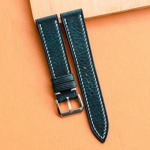 以下が含まれることがあります： 白いステッチとシルバーのバックルが付いた、濃い緑色の革製腕時計ストラップ。 ストラップは、背景に木の板があるオレンジ色の表面に表示されています。 腕時計ストラップは新品です。