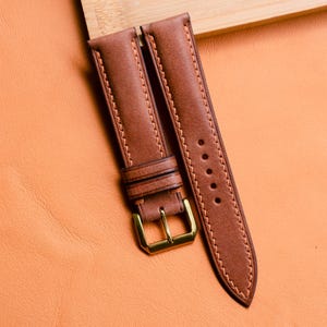 Pode incluir: Pulseira de relógio de couro marrom com fivela dourada. A pulseira tem um design costurado com uma linha marrom mais clara. A pulseira do relógio é mostrada em uma superfície laranja texturizada.