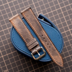 Peut inclure: Un bracelet de montre en cuir marron avec des coutures beige apparentes, deux passants en cuir et une boucle en métal argenté poli. Le bracelet repose sur un étui de montre en cuir bleu texturé, posé sur une surface à motif quadrillé.