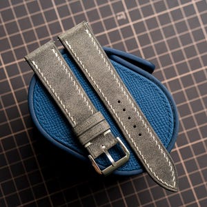 May include: A two-piece grey watch strap with a textured surface and white contrast stitching. The strap features a silver-toned metal buckle and multiple adjustment holes, resting on a blue textured oval-shaped watch display cushion.
