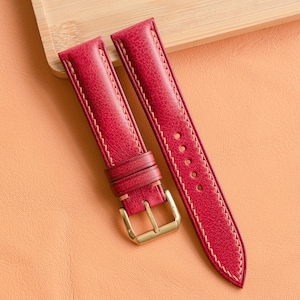 May include: Two red leather watch straps with white stitching and gold-coloured buckles. The straps are lying on a light orange surface, with a wooden block in the background.