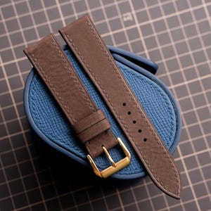 May include: A dark brown leather watch strap with visible stitching along the edges and a polished gold-tone metal buckle. The strap is composed of two pieces, one featuring a series of adjustment holes and the other with a fixed and a floating keeper.