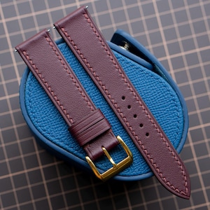 Könnte beinhalten: Ein tiefburgunderfarbenes Leder-Uhrenarmband mit passenden Nähten und einer polierten, goldfarbenen Metallschnalle, das auf einer strukturierten blauen Leder-Uhrenrolle liegt. Das Armband hat eine Reihe von Einstelllöchern und eine feste Lederschlaufe.
