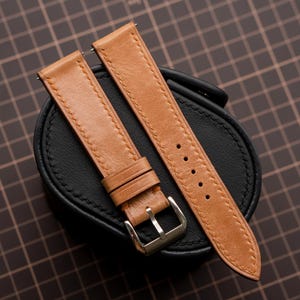 May include: A tan leather watch strap featuring visible stitching along the edges, two leather keepers, and a silver-toned metal buckle. The strap is displayed resting on a black, oval-shaped, textured watch holder.