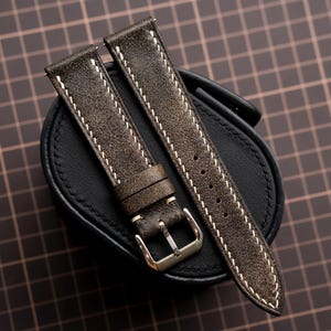Peut inclure: Un bracelet de montre texturé marron foncé avec des surpiqûres contrastantes blanches sur les bords. Le bracelet comprend deux passants en cuir et une boucle en métal argenté poli, posé sur un coussin de présentation noir.