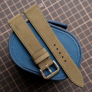 Op de afbeelding: Een tweedelige horlogeband van olijfgroen leer met ton-sur-ton stiksels, een zilverkleurige metalen gesp en een reeks gestanste gaten voor verstelling. De band ligt op een gestructureerd, ovaalvormig blauw opbergdoosje voor horloges.