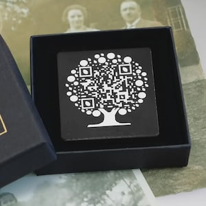 Puede incluir: Una caja negra con letras doradas que dicen "memo rio". Dentro de la caja hay un cuadrado negro con un código QR blanco en forma de árbol. Una rosa blanca y una fotografía vintage en el fondo.