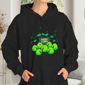 Puede incluir: Sudadera con capucha negra con un gráfico de personajes de dibujos animados verdes y el texto "HARD CARRY". El diseño incluye notas musicales y la palabra "HA" repetida. La sudadera tiene un bolsillo delantero y una capucha con cordón.