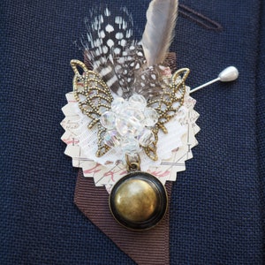 May include: A unique lapel pin featuring a decorative butterfly design with bronze-colored filigree wings, clear beads, and a dangling bronze pendant. The pin includes a cluster of feathers in shades of gray, brown, and white, and is attached with a pin.