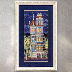 Puede incluir: Obra de arte en punto de cruz enmarcada de una casa de estilo victoriano por la noche. La casa es de varios pisos con detalles en rosa, azul y marrón, ventanas iluminadas y un cielo estrellado de fondo. La obra está enmarcada en un marco blanco ornamentado.