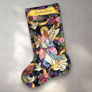 Puede incluir: Un calcetín navideño con un ángel tocando el arpa, rodeado de rosas y palomas. El calcetín tiene un fondo negro con el nombre "Samantha" en una pancarta amarilla. El ángel viste un vestido blanco, rosa y azul.
