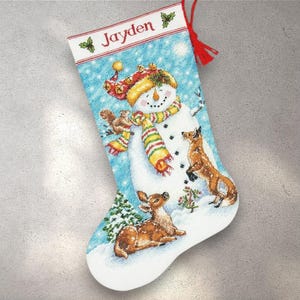 Puede incluir: Calcetín navideño con un muñeco de nieve, un ciervo y un zorro en una escena nevada. El calcetín es azul claro con el nombre "Jayden" en la parte superior. El muñeco de nieve lleva un sombrero y una bufanda. Una borla roja cuelga de la parte superior.