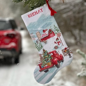 Op de afbeelding: Witte kerstsok met de naam "RIENAT" in het rood. De sok toont een winterse scène met een rode vrachtwagen met een kerstboom, een sneeuwpop en een schuur. De woorden "MERRY CHRISTMAS" zijn ook zichtbaar.