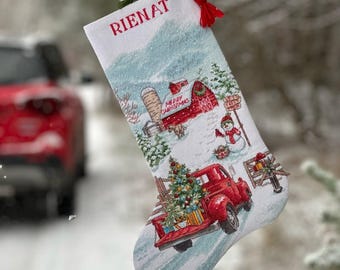 May include: White Christmas stocking with the name "RIENAT" in red. The stocking features a winter scene with a red truck carrying a Christmas tree, a snowman, and a barn. The words "MERRY CHRISTMAS" are also visible.