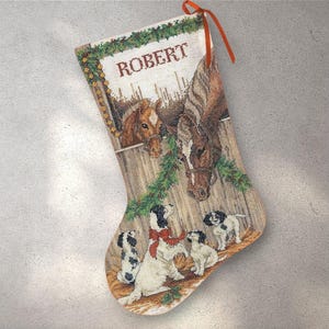 Può includere: Calza di Natale a punto croce con cavalli e cuccioli. La calza ha il nome "ROBERT" in alto. Il design include cavalli in una stalla, cuccioli e un nastro rosso per appenderla. La tavolozza dei colori comprende marrone, bianco e verde.