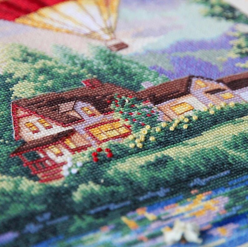 Puede incluir: Un paisaje de punto de cruz representa una casa con tejado marr&oacute;n, detalles rojos y ventanas iluminadas. Un globo aerost&aacute;tico flota en el cielo sobre la casa, rodeado de &aacute;rboles verdes y un cielo azul y morado. La obra de arte es detallada y colorida.