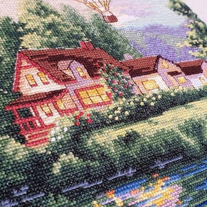 Puede incluir: Bordado de punto de cruz que representa una pintoresca escena de pueblo con casas, &aacute;rboles y un globo aerost&aacute;tico. Las casas tienen tejados marrones, detalles en rojo y ventanas iluminadas. La escena est&aacute; llena de follaje verde y un r&iacute;o azul.