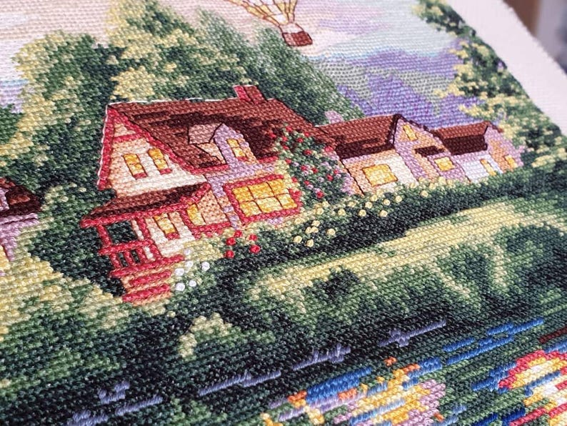 Puede incluir: Bordado de punto de cruz que representa una pintoresca escena de pueblo con casas, &aacute;rboles y un globo aerost&aacute;tico. Las casas tienen tejados marrones, detalles en rojo y ventanas iluminadas. La escena est&aacute; llena de follaje verde y un r&iacute;o azul.