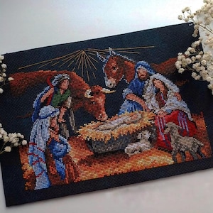 Puede incluir: Un bordado de punto de cruz de la Natividad, que representa a María, José y el niño Jesús en un pesebre. La obra de arte incluye un burro, un buey y ovejas, con un fondo azul oscuro y tonos cálidos y terrosos.