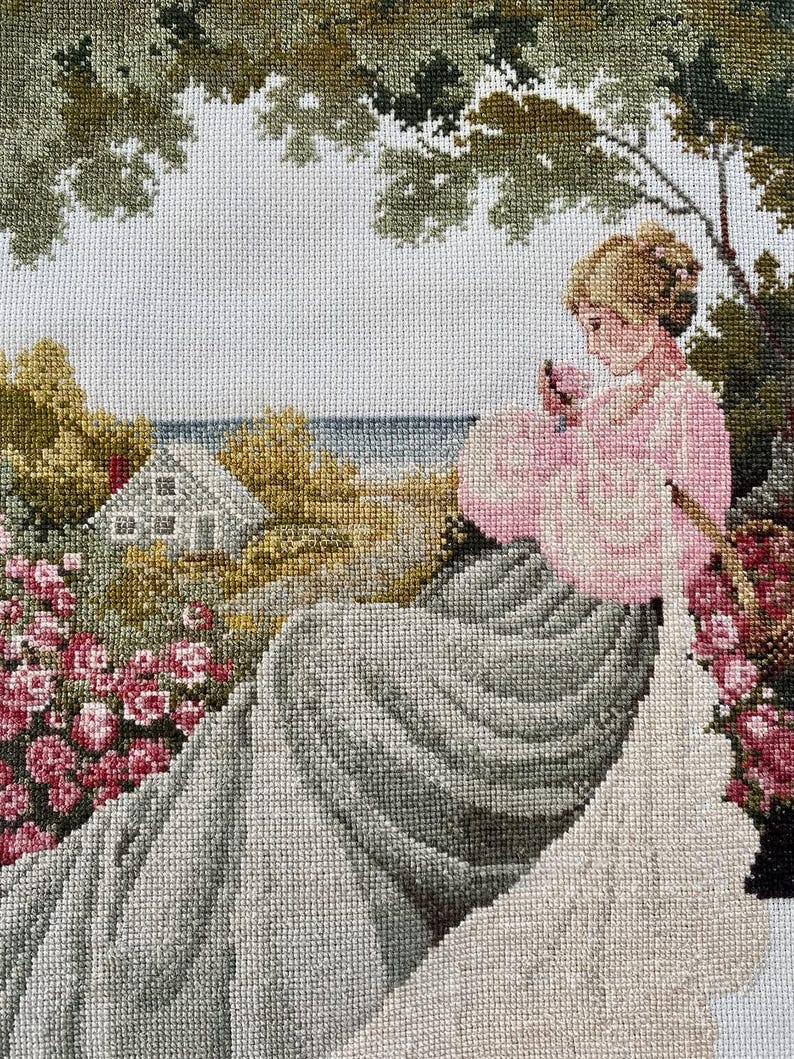 May include: A detailed needlepoint tapestry depicting a woman in a pink dress seated in a garden. The scene includes a house, flowers, and trees, with a coastal view in the background. The artwork is rendered in a cross-stitch style.