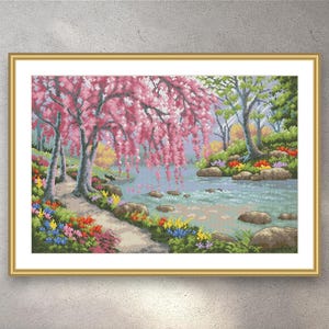 May include: Cross-stitch landscape art depicting a tranquil river scene. Pink blossom trees cascade over the water, with colourful flowers lining the banks. The artwork is framed in a gold-coloured frame, creating a peaceful and decorative piece.
