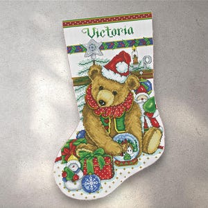 May include: Embroidered Christmas stocking with a teddy bear in a Santa hat and red collar. The stocking is decorated with gifts, a snowman, and a snow globe. The name "Victoria" is embroidered at the top.