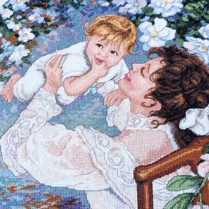 May include: Cross-stitch of a mother and child. The mother, with brown curly hair, is wearing a white dress with lace details. The child, with blonde hair, is wearing a white outfit. The background features blue and white flowers.