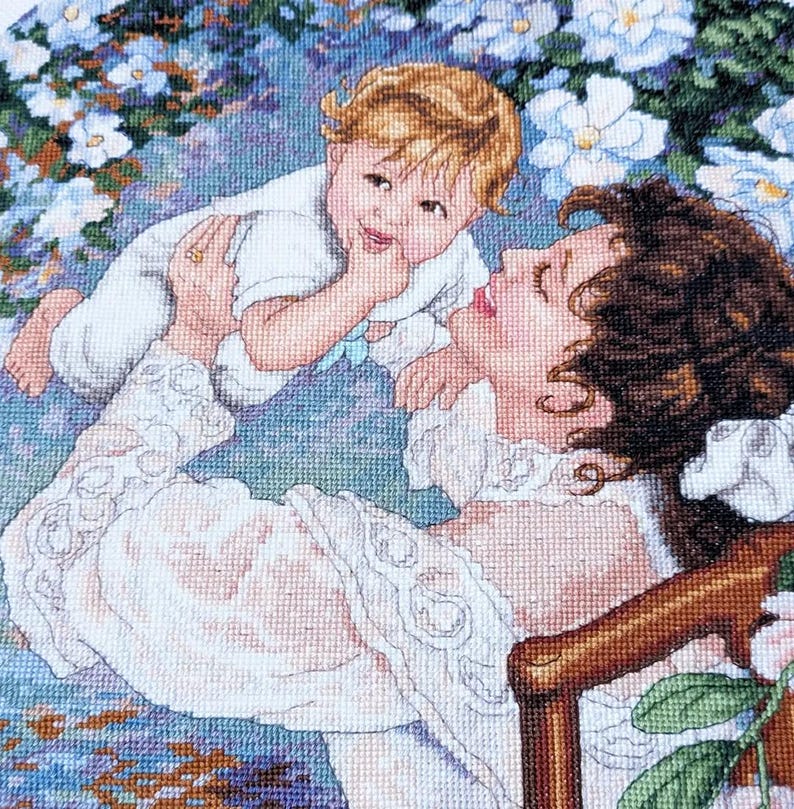May include: Cross-stitch of a mother and child. The mother, with brown curly hair, is wearing a white dress with lace details. The child, with blonde hair, is wearing a white outfit. The background features blue and white flowers.
