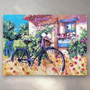 May include: Cross-stitch artwork showing a vintage bicycle with a basket, parked outside a cottage. The scene is filled with flowers and greenery, with a blue sky. The bicycle is black, and the basket has a pink ribbon.