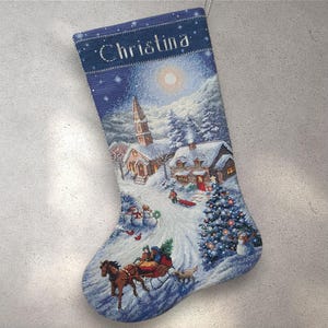 Op de afbeelding: Een kerstsok met een winterdorpsscène. De sok is blauw met de naam "Christina" bovenaan. De scène toont een besneeuwd dorp met een kerk, huizen, een paardenslee en een versierde kerstboom.