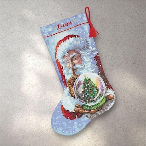 以下が含まれることがあります： クリスマスストッキング。サンタクロースがクリスマスツリーの入ったスノーグローブを持っています。ストッキングは水色で雪の結晶模様、名前「Liam」は赤色。赤いタッセルが上部に付いています。