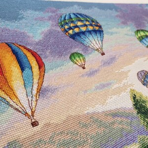 Puede incluir: Un bordado de punto de cruz que representa globos aerost&aacute;ticos de colores flotando en un cielo nublado. Los globos son de varios colores, incluyendo rojo, amarillo, azul y verde. El fondo presenta un degradado de azules, morados y naranjas.