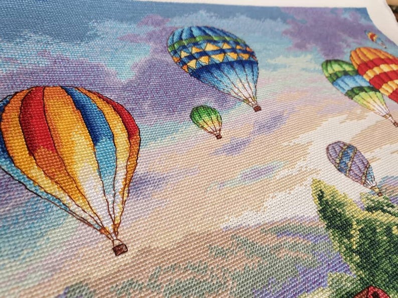 Puede incluir: Un bordado de punto de cruz que representa globos aerost&aacute;ticos de colores flotando en un cielo nublado. Los globos son de varios colores, incluyendo rojo, amarillo, azul y verde. El fondo presenta un degradado de azules, morados y naranjas.
