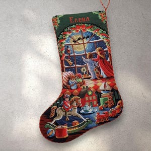 Puede incluir: Un calcetín navideño con un diseño de tapiz detallado. La parte superior es verde con el nombre "Elena". La imagen principal representa una escena invernal con niños, juguetes y una luna llena. El calcetín es rojo y presenta una variedad de juguetes.